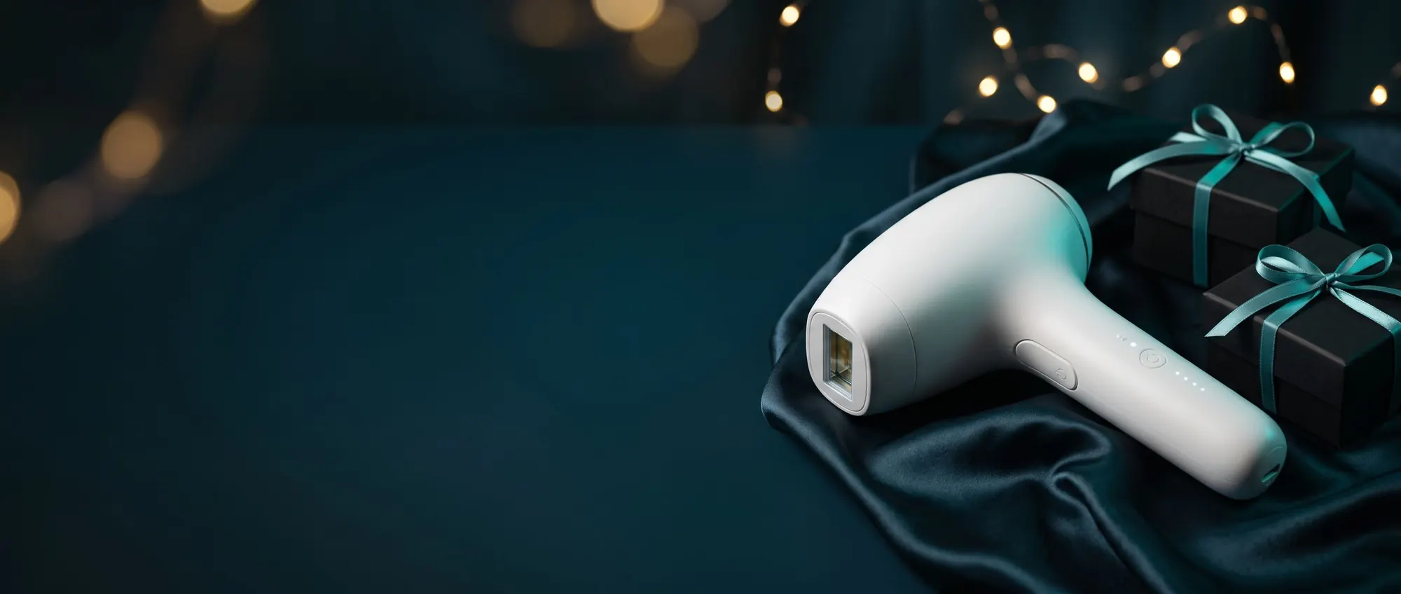 Epilatore a luce pulsata bianco su un drappo di raso accanto a pacchi regalo neri con nastro verde acqua